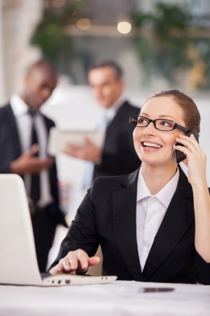 Businesswoman on the phone. Cheerful young woman in formalwear sitting at the table and talking on the mobile phone while two colleagues standing on backgroundの写真素材