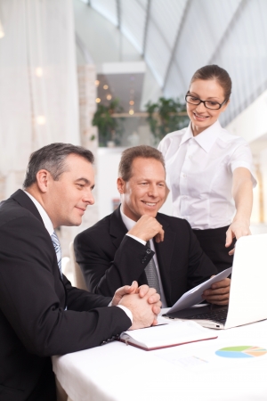 It was a good deal. Two cheerful men in formalwear sitting at the table while woman in white shirt standing close to them and pointing computer monitorの写真素材