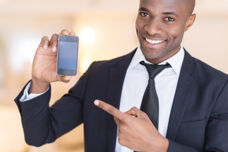 Businessman with mobile phone. Cheerful young African man in formalwear holding a mobile phone and pointing itの写真素材