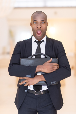 Frustrated businessman. Shocked young African man in full suit holding a bag in his hands and keeping mouth open while looking at cameraの写真素材