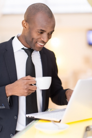 Business on the go. Cheerful young African businessman using his laptop while standing at the bar の写真素材