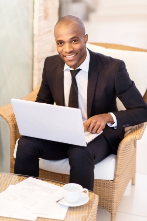 Businessman with laptop. Cheerful young African businessman using his laptop and smiling at camera while sitting at the restaurant の写真素材