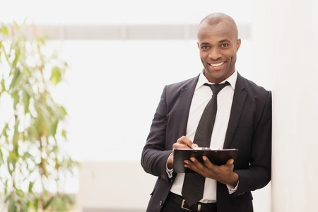 Businessman with note pad. Cheerful young African businessman writing something in his note pad and smiling at cameraの写真素材