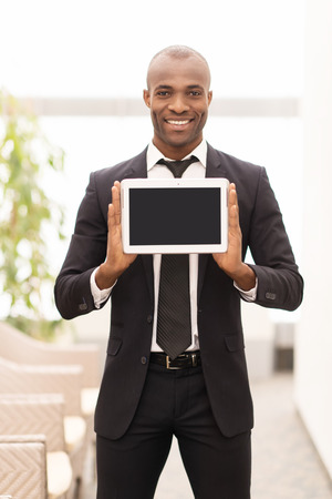 Join a digital age. Cheerful young African man in formalwear showing a screen of digital tablet and smiling at cameraの写真素材
