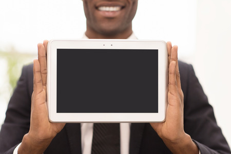 Copy space on his tablet. Cropped image of cheerful young African man in formalwear showing a screen of digital tablet and smiling at cameraの写真素材