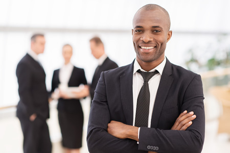 Confident businessman. Cheerful young African man in formalwear keeping arms crossed and smiling while his colleagues standing on backgroundの写真素材