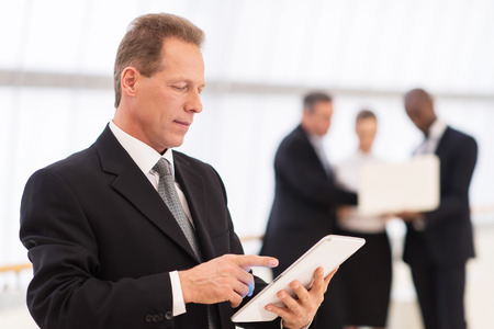 Businessman with digital tablet.  Thoughtful mature man in formalwear using digital tablet while his colleagues standing on backgroundの写真素材