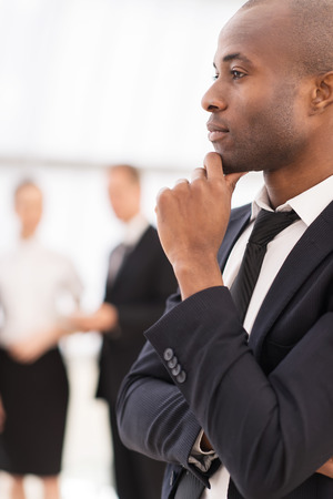 Thoughtful businessman.  Thoughtful young African man in formalwear holding hand on chin and looking away while his colleagues  standing on backgroundの写真素材