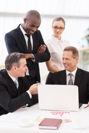 Business team at meeting. Two cheerful mature man in formalwear sitting at the table and communicating while two another people standing close to them の写真素材