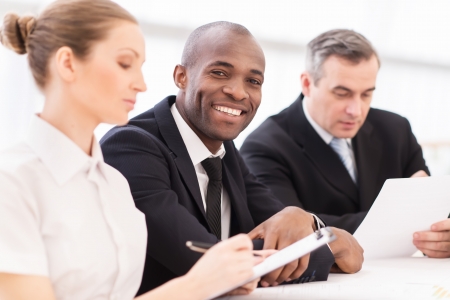 Man on business meeting. Cheerful young African man in formalwear looking at camera and smiling at camera while sitting together with his colleaguesの写真素材