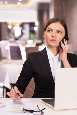 Confident businesswoman. Thoughtful young woman in formalwear talking on the phone and looking away while sitting at the restaurantの写真素材