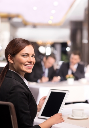 Businesswoman with digital tablet. Rear view of cheerful young woman in formalwear working on digital tablet and looking over shoulder while sitting at the restaurantの写真素材