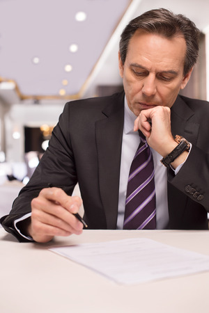 Signing a contract. Thoughtful mature man in formalwear signing a document and holding hand on chinの写真素材