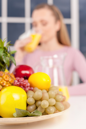 Healthy food and drink. Beautiful young woman drinking orange juice while plate with fruits laying on foreground の写真素材