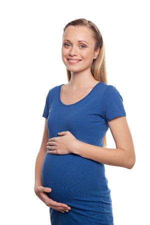 Waiting for a baby. Cheerful young pregnant woman touching her abdomen and looking at camera while standing isolated on whiteの写真素材
