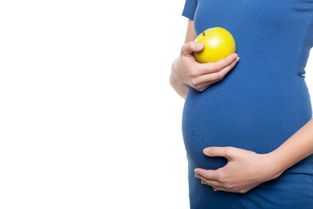 Waiting for a baby. Cropped image young pregnant woman touching her abdomen and holding apple while standing isolated on whiteの写真素材
