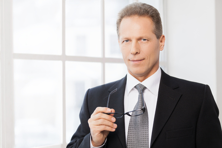 Smart and confident. Portrait of confident mature man in formalwear holding glasses and smiling at camera while standing near window の写真素材