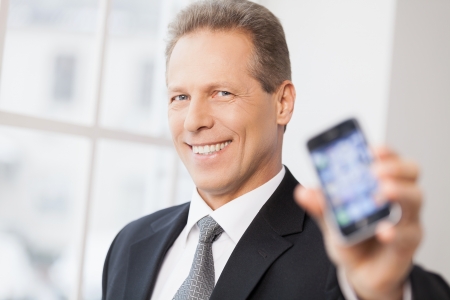 Businessman with mobile phone. Portrait of cheerful senior man in shirt and tie showing his mobile phone to camera and smiling の写真素材