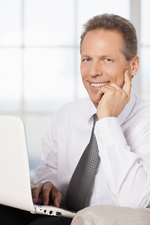 Businessman with laptop. Cheerful mature man in shirt and tie using computer and holding hand on chinの写真素材