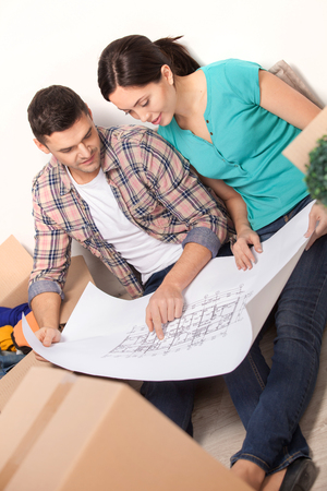 Planning a house improvement. Top view of young couple sitting on the floor and looking at the blueprint while cardboard boxes laying around them の写真素材