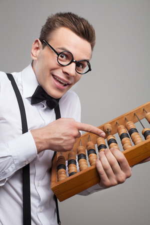 cheerful young man in bow tie and suspenders holding abacus and pointing it while standing against grey backgroundの写真素材