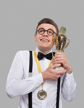 Cheerful young man in bow tie holding a trophy and looking up while standing against grey background の写真素材