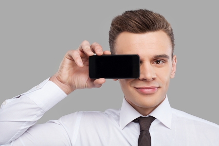 Handsome young man in shirt and tie holding mobile phone in front of the eye and smiling while standing against grey backgroundの写真素材