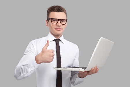 Man with laptop. Handsome young man in shirt and tie holding a laptop and gesturing while standing against grey backgroundの写真素材