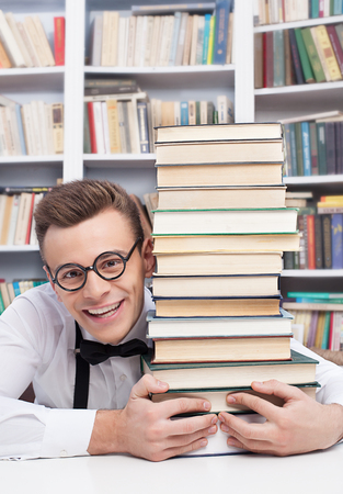 I love studying. Cheerful young man in shirt and bow tie sitting at the table in library and hugging a book stack の写真素材