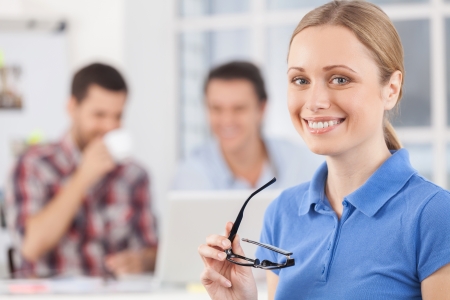 Happy to be a part of the team. Cheerful young woman holding glasses and smiling at camera while her colleagues working の写真素材