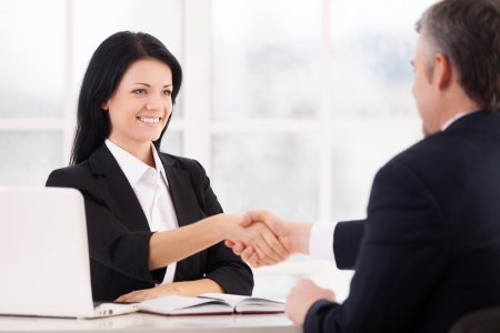 Sealing a deal. Two cheerful business people handshaking and smiling while sitting face to face at the tableの写真素材
