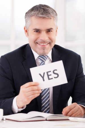 Approved! Cheerful grey hair man in formalwear holding a paper with yes sign on it while sitting at her working placeの写真素材