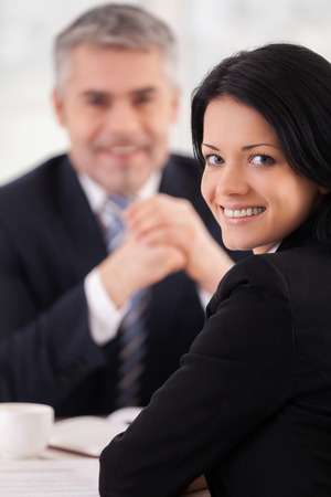 Woman on interview. Cheerful young woman in formal wear looking over shoulder and smiling while mature man in suit sitting の写真素材