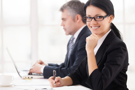 Business people at work. Cheerful young businesswoman sitting at the table and smiling at camera while mature man working on laptop on backgroundの写真素材