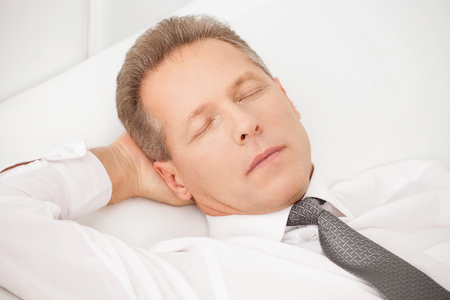 Sleeping businessman. Tired grey hair man in shirt and tie lying on bed and keeping eyes closedの写真素材