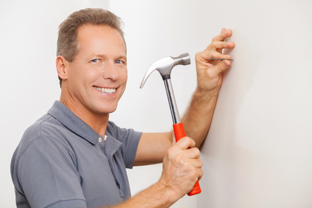 Cheerful handyman. Cheerful grey hair repairman hammering a nail and smiling at cameraの写真素材