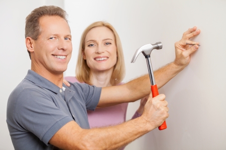 Repairing home together. Cheerful grey hair man hammering a nail and smiling while his wife standing close to himの写真素材
