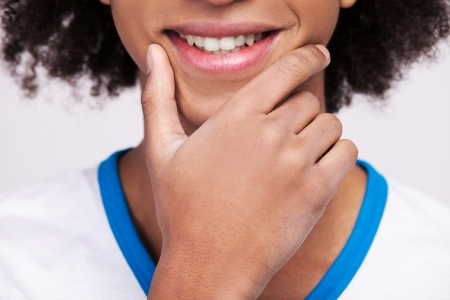 Cheerful teenager. Cropped image of cheerful African teenager smiling and holding hand on chin while standing isolated on grey backgroundの写真素材