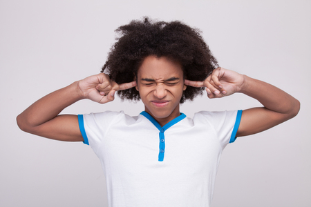 Hear no evil. Funky African teenager making a face and covering ears by fingers while standing isolated on grey の写真素材