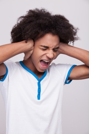 Stop it! Furious African teenager shouting and covering ears by hands while standing against grey の写真素材