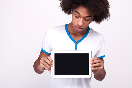 Looking at copy space. Curious African teenager on touchscreen of digital tablet while standing isolated on grey の写真素材