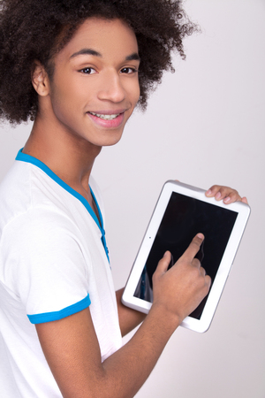 Working on digital tablet. Rear view of cheerful African teenager working on digital tablet and looking over shoulder while standing isolated on grey の写真素材