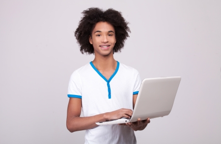 Teenager with laptop. Cheerful African teenager working on laptop and smiling at camera while standing isolated on grey の写真素材