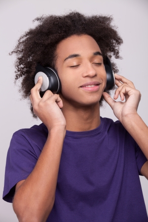 Listening to his favorite music. Cheerful African teenager in headphones listening to the music and grimacing while standing isolated on grey の写真素材