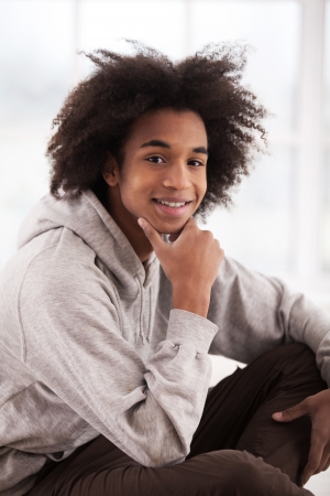 Young and carefree. Cheerful teenage African boy sitting on the floor and holding hand on chin の写真素材