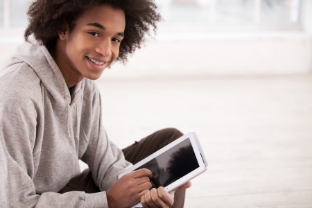 Surfing web at free time. Cheerful African teenager working on digital tablet and while sitting on the floorの写真素材