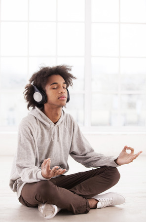 Chill time. Teenage African boy in headphones listening to the music and keeping eyes closed while sitting in lotus position on the floorの写真素材