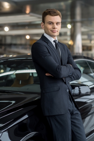 Confident in his choice. Confident man in formalwear leaning at the car and looking at camera while standing at car dealership