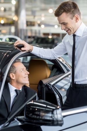 Let me show you all the features. Confident mature man ion formalwear sitting at the front seat of the car and talking to the car salesman standing near himの写真素材