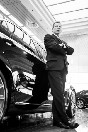 Only luxury cars. Black and white image of full length confident grey hair man in formalwear leaning at the car and looking awayの写真素材